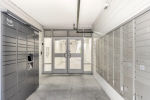 Interior hallway, cluster mailbox units, gray amazon hub drop box across from mailbox units, White walls.